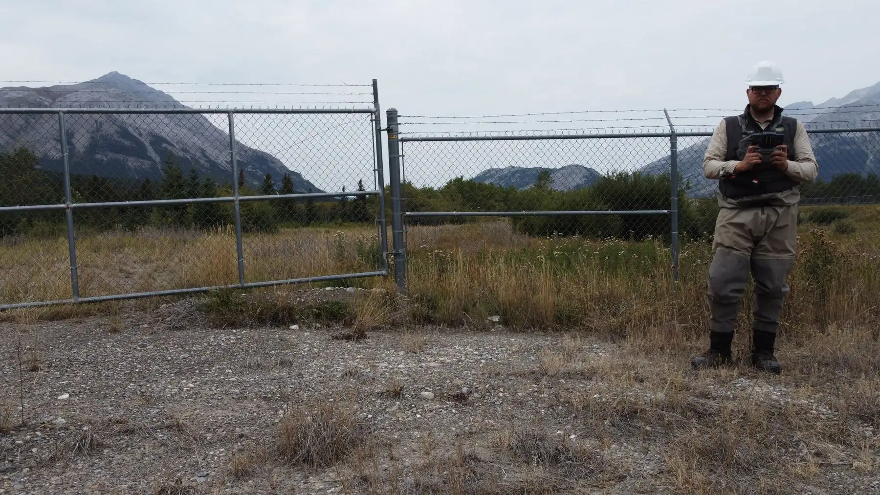 Kirby in waders with a clipboard beside a chain-link fence, mountains behind