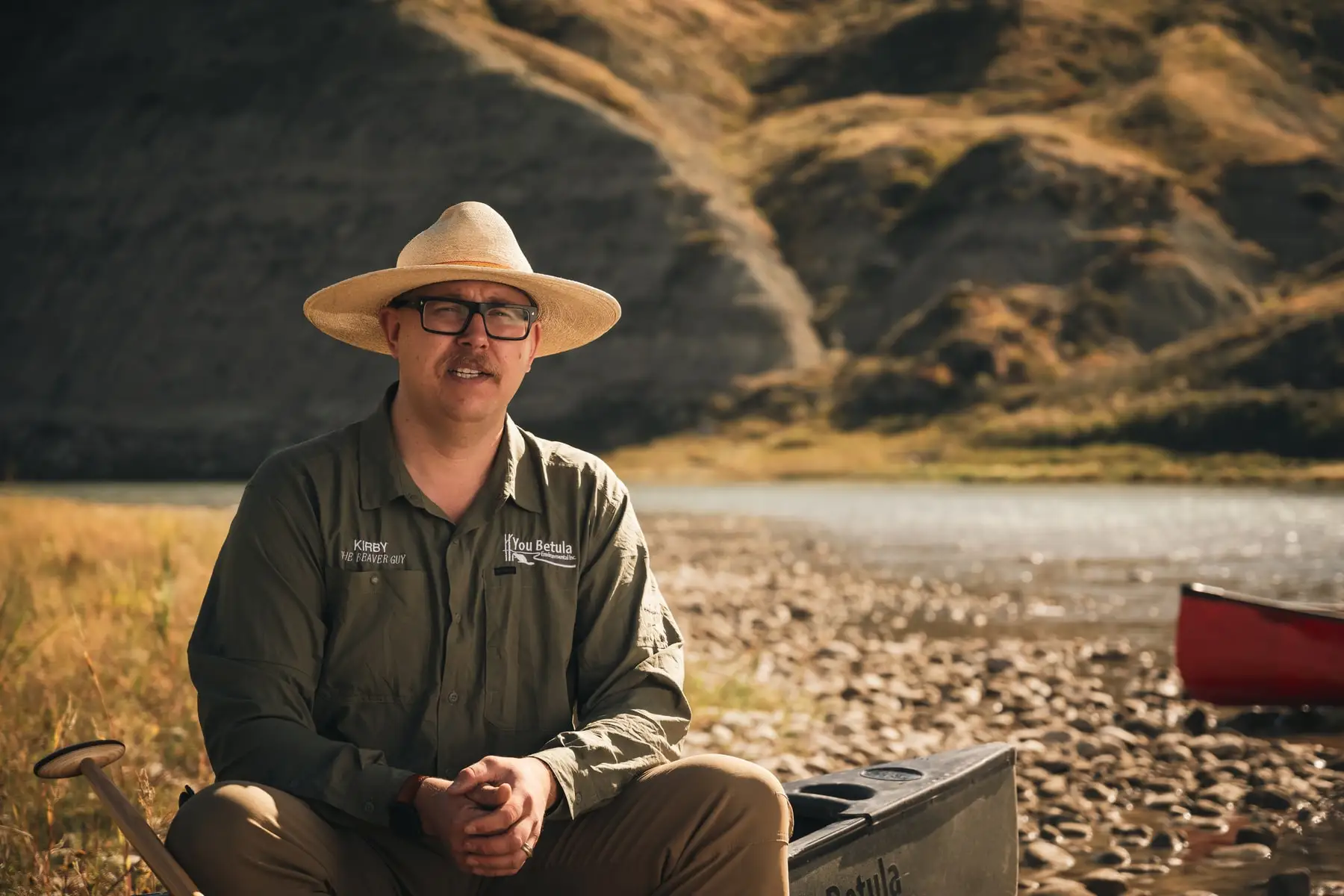 Kirby England seated beside a red canoe on a gravel bar in the riverbottom